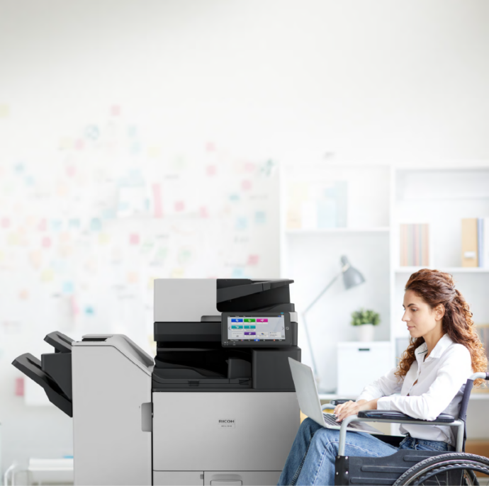 Professional using a laptop next to a Ricoh multifunction printer, highlighting Woodhull’s accessible and inclusive office technology solutions for diverse work environments.