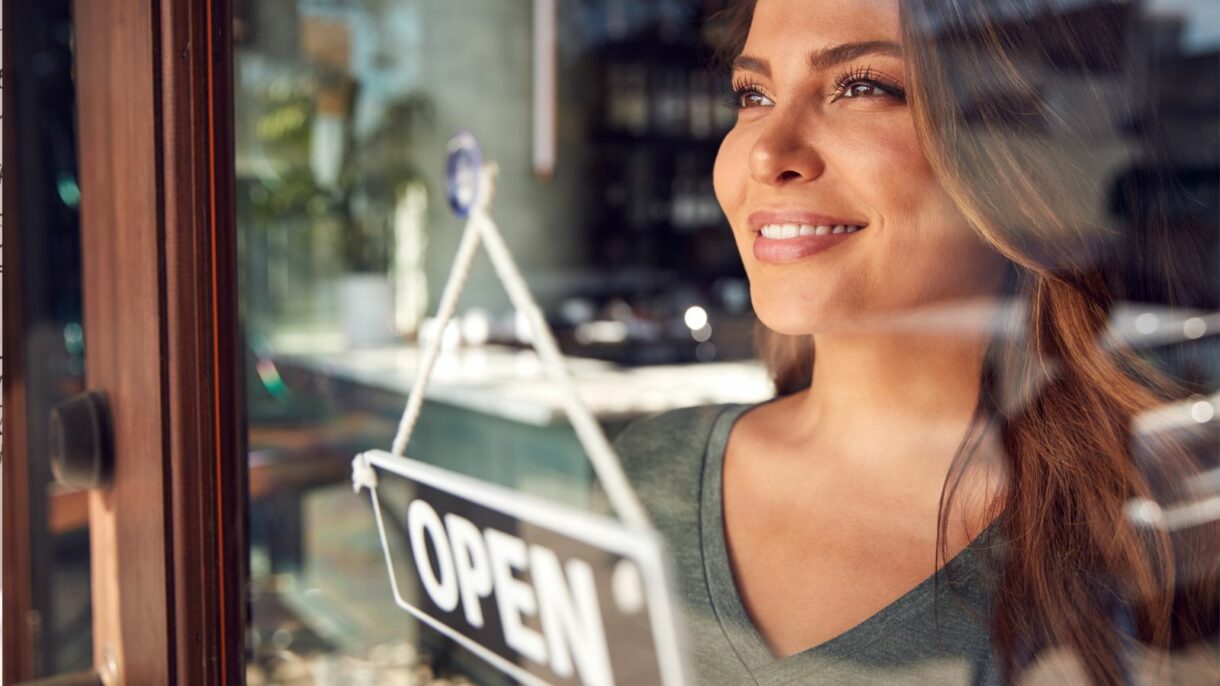 Smiling woman looking out of a shop window with an 'Open' sign, symbolizing Woodhull’s support for women-owned businesses as a WBENC-certified company.