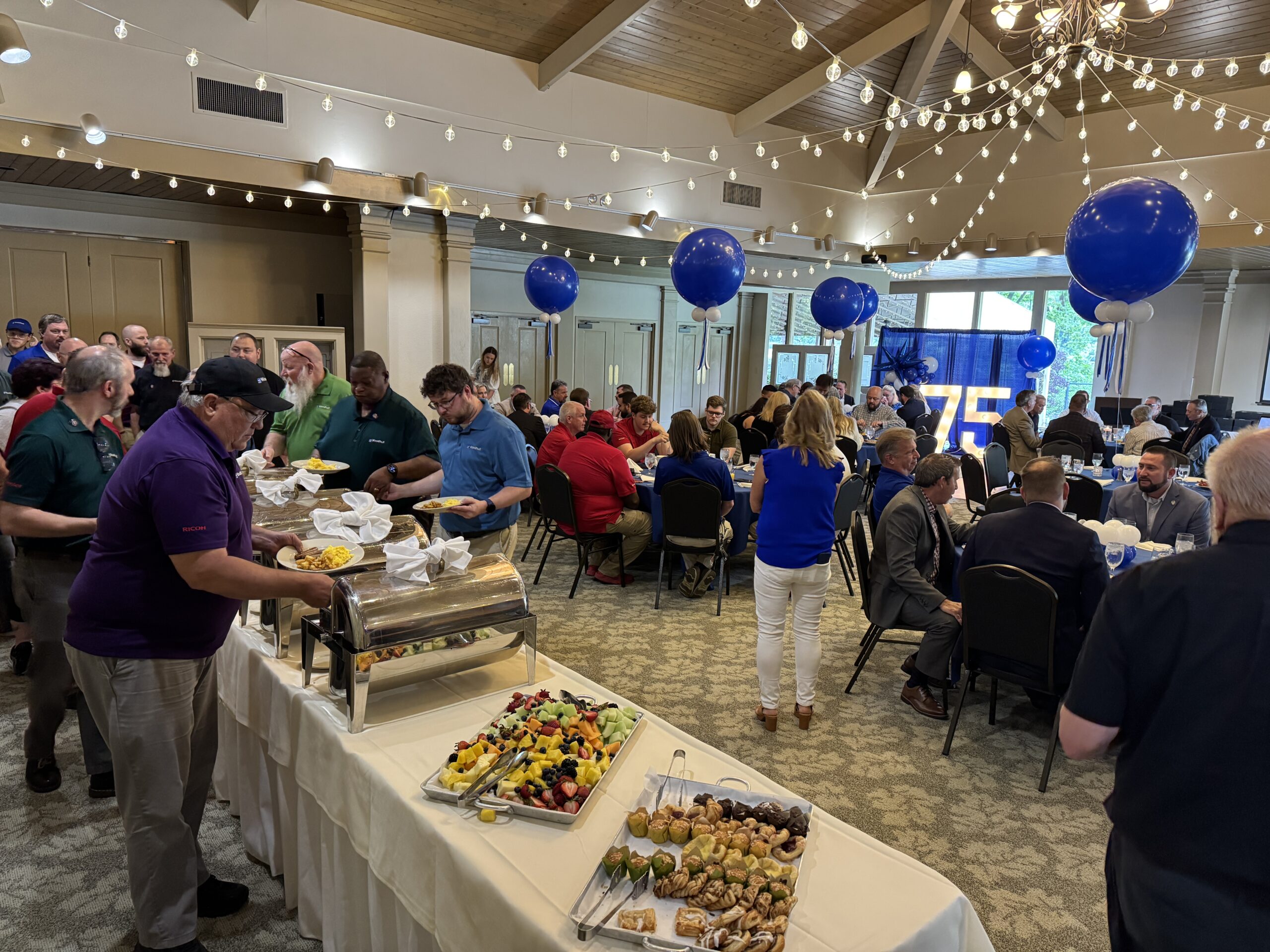 Woodhull employees and guests gather in a decorated banquet hall with string lights and blue balloons to celebrate the company’s 75th anniversary as a 100% employee-owned ESOP. Attendees are seen enjoying a buffet-style meal with fruit and pastries, while others are seated at round tables in front of a large illuminated ‘75’ display.