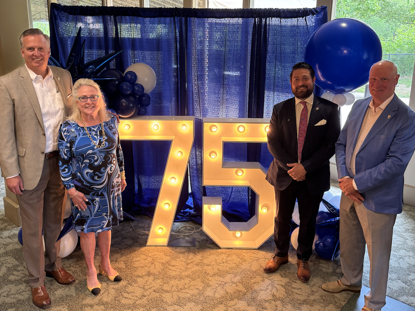 Four professionally dressed individuals stand in front of a large illuminated '75' sign at an indoor celebration, with blue and white balloons and a navy curtain backdrop, marking Woodhull’s 75th anniversary as a 100% employee-owned ESOP company.